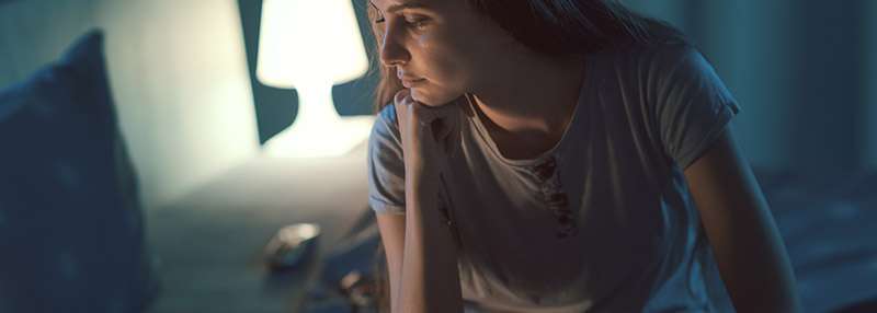 Eine nachdenkliche Frau sitzt im Dunkeln auf einem Bett, neben ihr leuchtet eine Lampe.