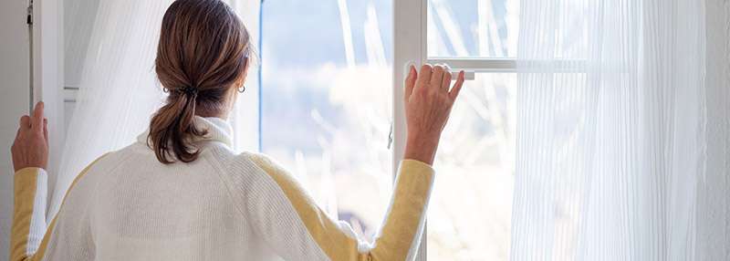 Eine Person zieht Vorhänge zurück und schaut aus einem Fenster in eine helle, natürliche Umgebung.
