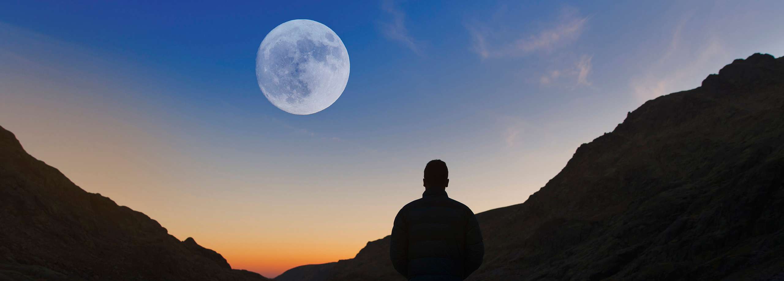 Ein Mann steht in den Bergen bei Sonnenaufgang während der Vollmond am Himmel sichtbar ist