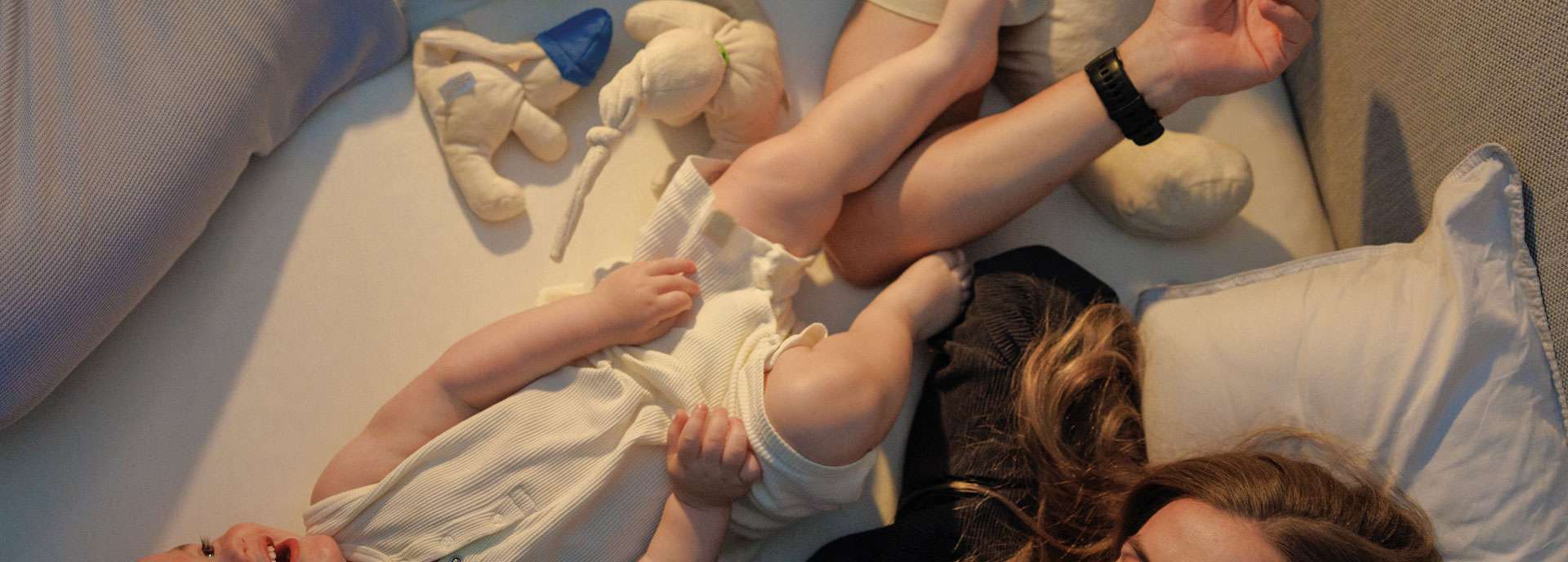 Familie entspannt im Bett mit einem Kleinkind, umgeben von Kissen und einer gemütlichen Atmosphäre.