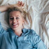 Lächelnde Frau in blauer Schlafanzugoberteil liegt entspannt auf einem Bett mit weißen Bettlaken.