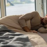 Ein älterer Mann schläft entspannt auf einem Bett mit Kissen, umgeben von einer ruhigen Landschaft.