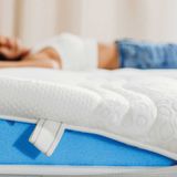Mann öffnet den Bezug einer Wenatex Matratze Comfort Plus, während eine Frau im Hintergrund entspannt liegt.