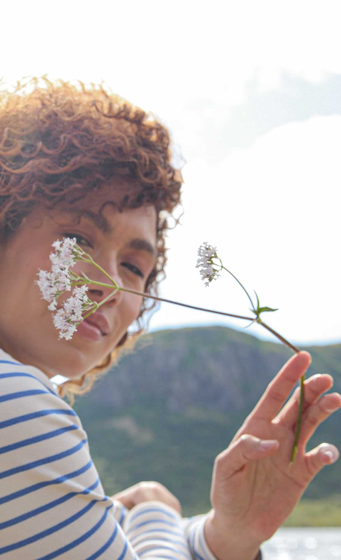 Woman with curly hair holds a flower in front of her face, with a green landscape in the background.