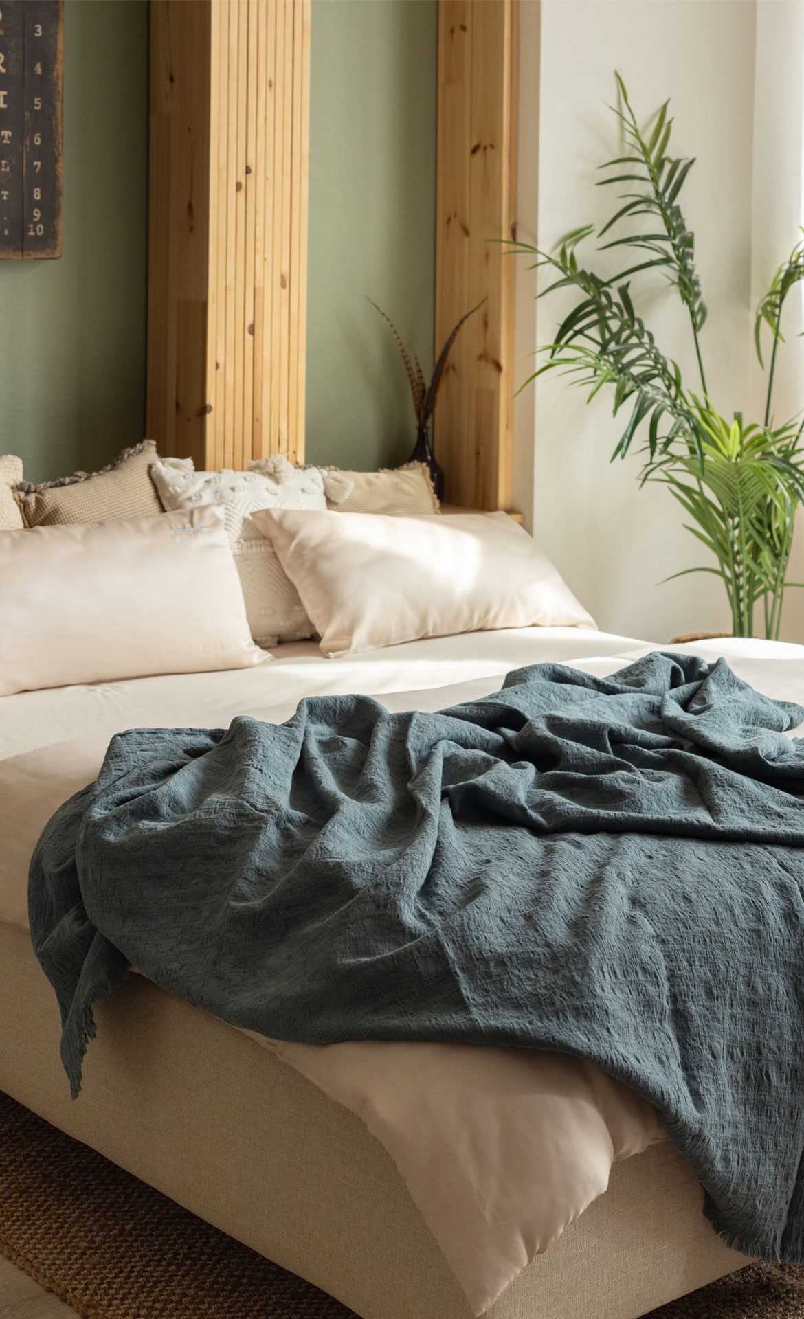 Bett mit hellen Kissen und einer blauen Decke, umgeben von Pflanzen und Holzdetails im Hintergrund.
