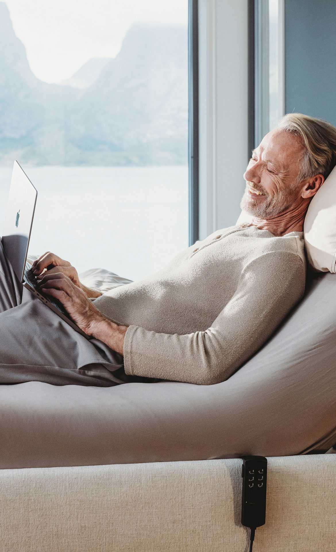 Man relaxing on a bed with a laptop, surrounded by natural light and scenic views.
