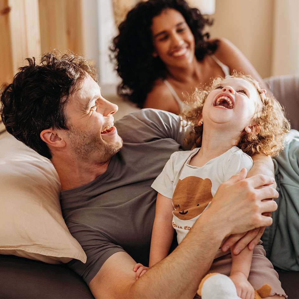 Eine lachende Familie entspannt auf einem Sofa, ein Elternteil umarmt ein fröhliches Kind.