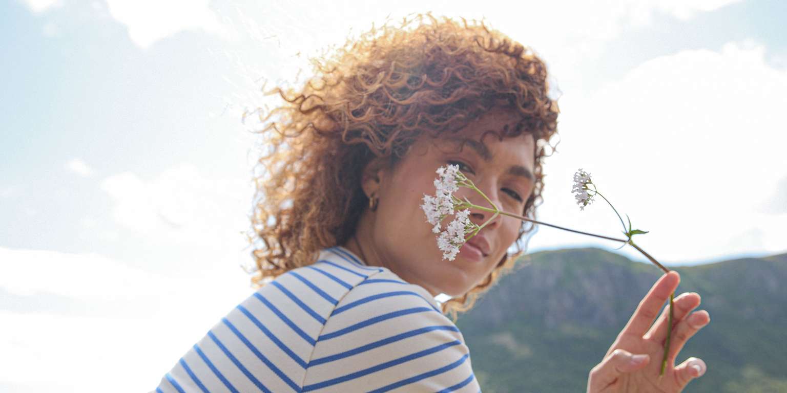 A woman with curly hair holds a flower in front of her face, surrounded by a natural landscape.