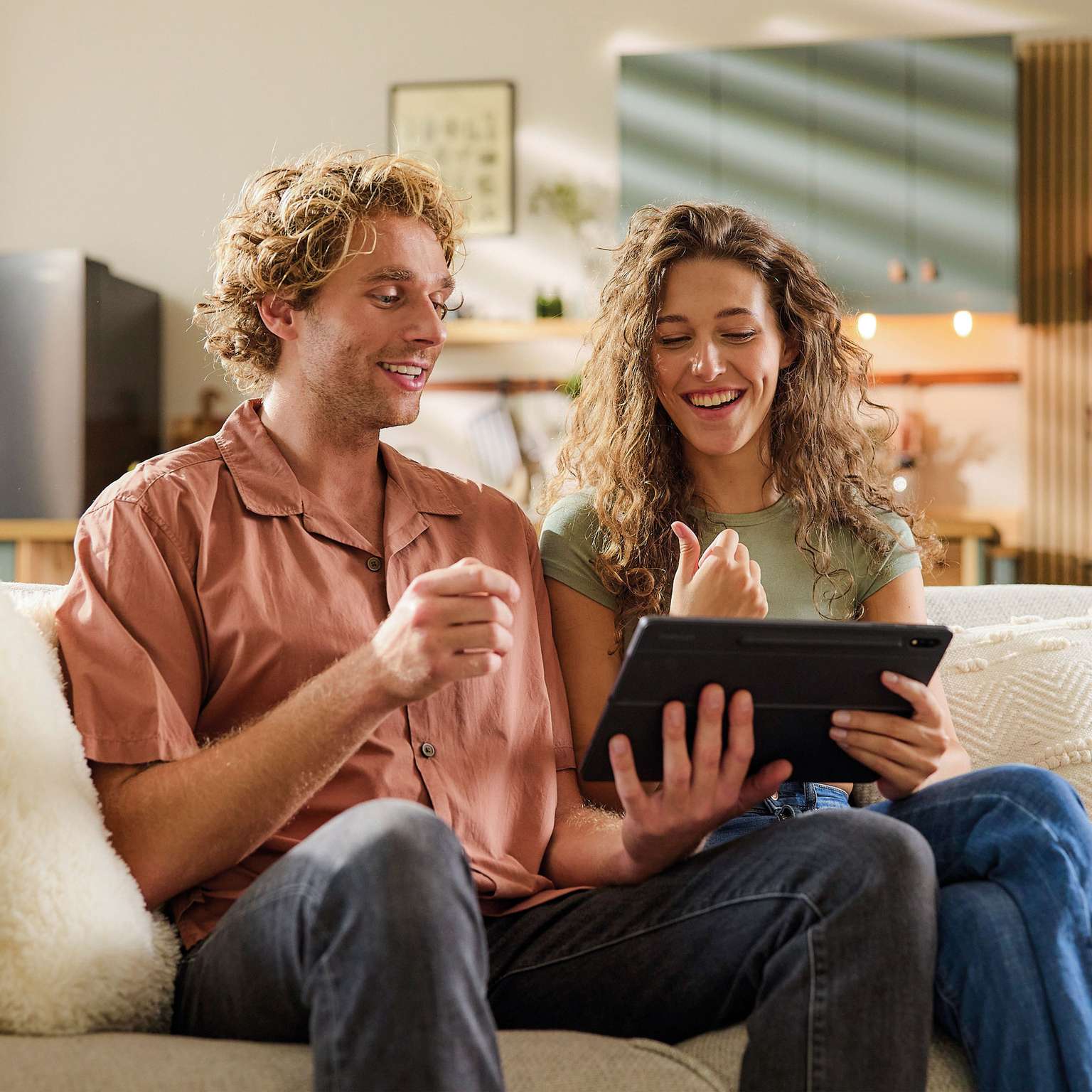 Couple sitting on a couch, smiling and discussing while looking at a tablet together.