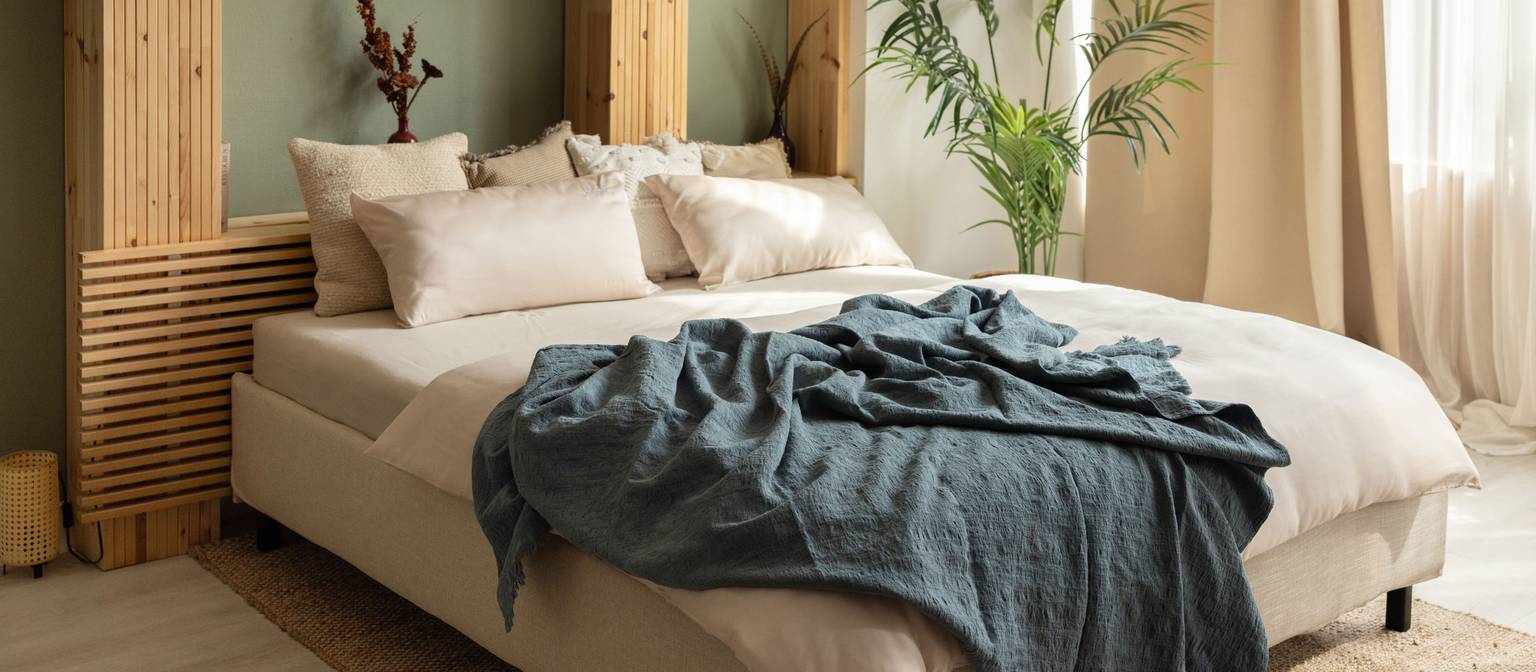 Cozy bedroom with bed, pillows, blanket, and plants in the background. Bright, inviting atmosphere.