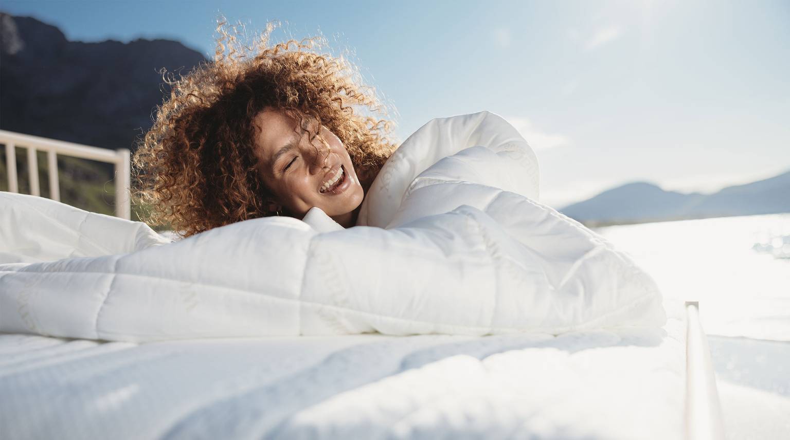 Woman with curly hair happily snuggled in a comforter on a bed.