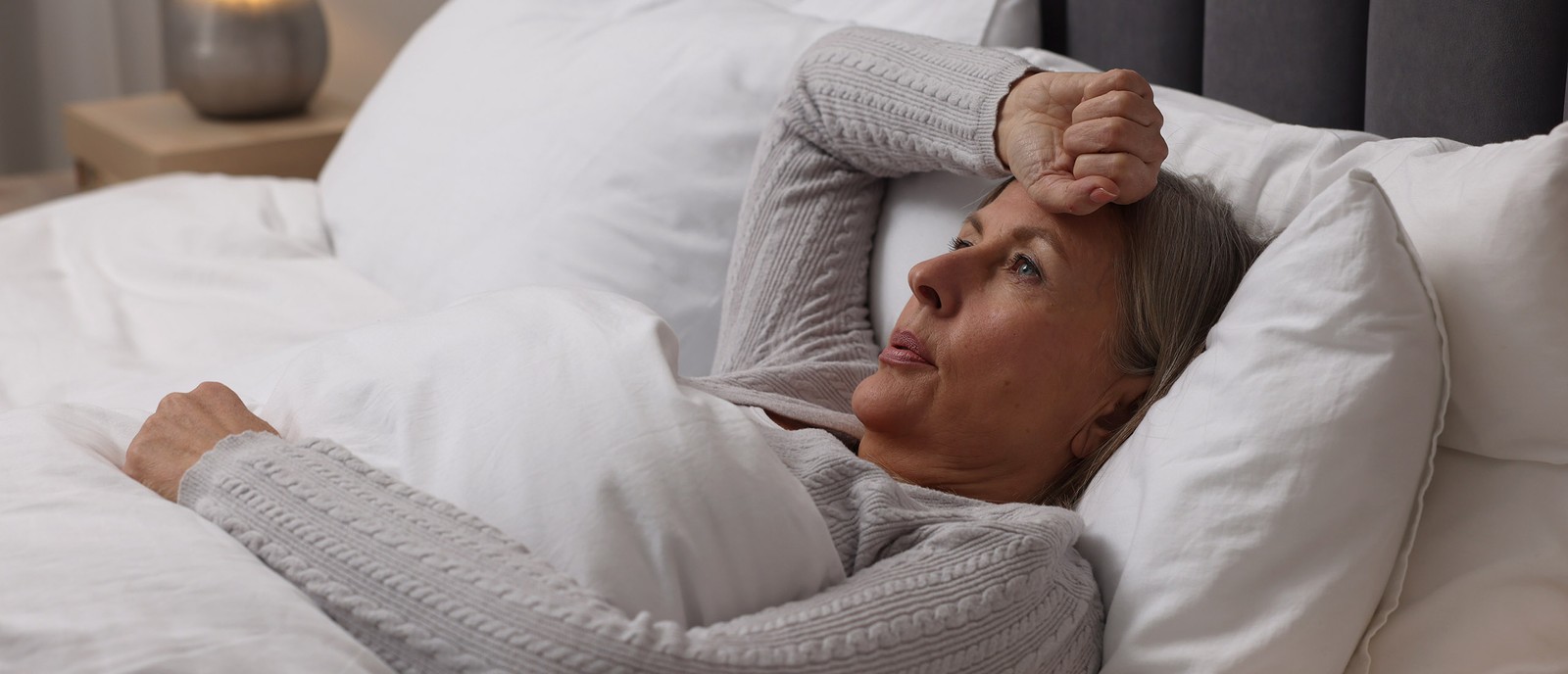 Una donna pensierosa è sdraiata a letto, sostenendo la testa con una mano, circondata da cuscini e coperte bianche.