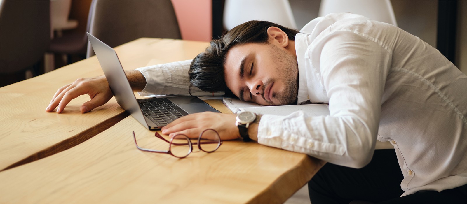 Mann schläft mit Kopf auf dem Tisch neben einem Laptop und einer Brille in einem modernen Raum.