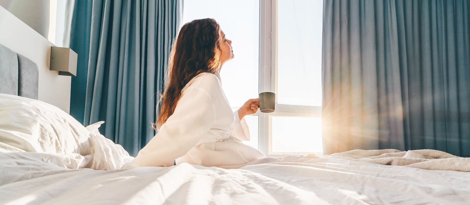 Frau in einem Bademantel sitzt auf dem Bett und genießt einen Kaffee bei Sonnenaufgang.