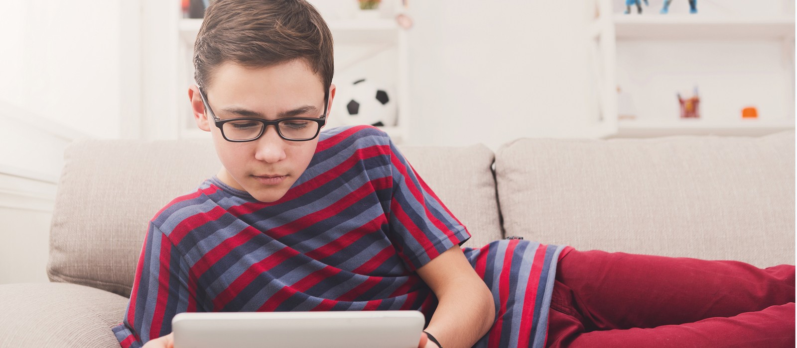Ragazzo con occhiali che usa un tablet su un divano, circondato da un ambiente luminoso.