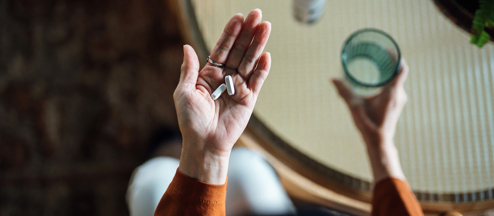Hände halten zwei Kapseln, während eine andere Hand ein Glas Wasser hält, auf einem Tisch.