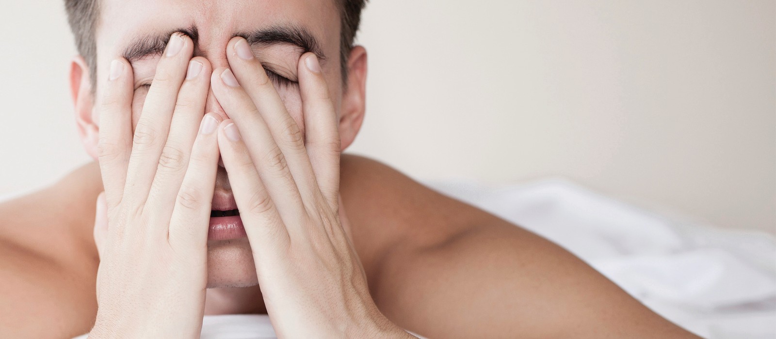 Mann liegt im Bett und hält sich frustriert die Hände vor das Gesicht, umgeben von Bettwaren.