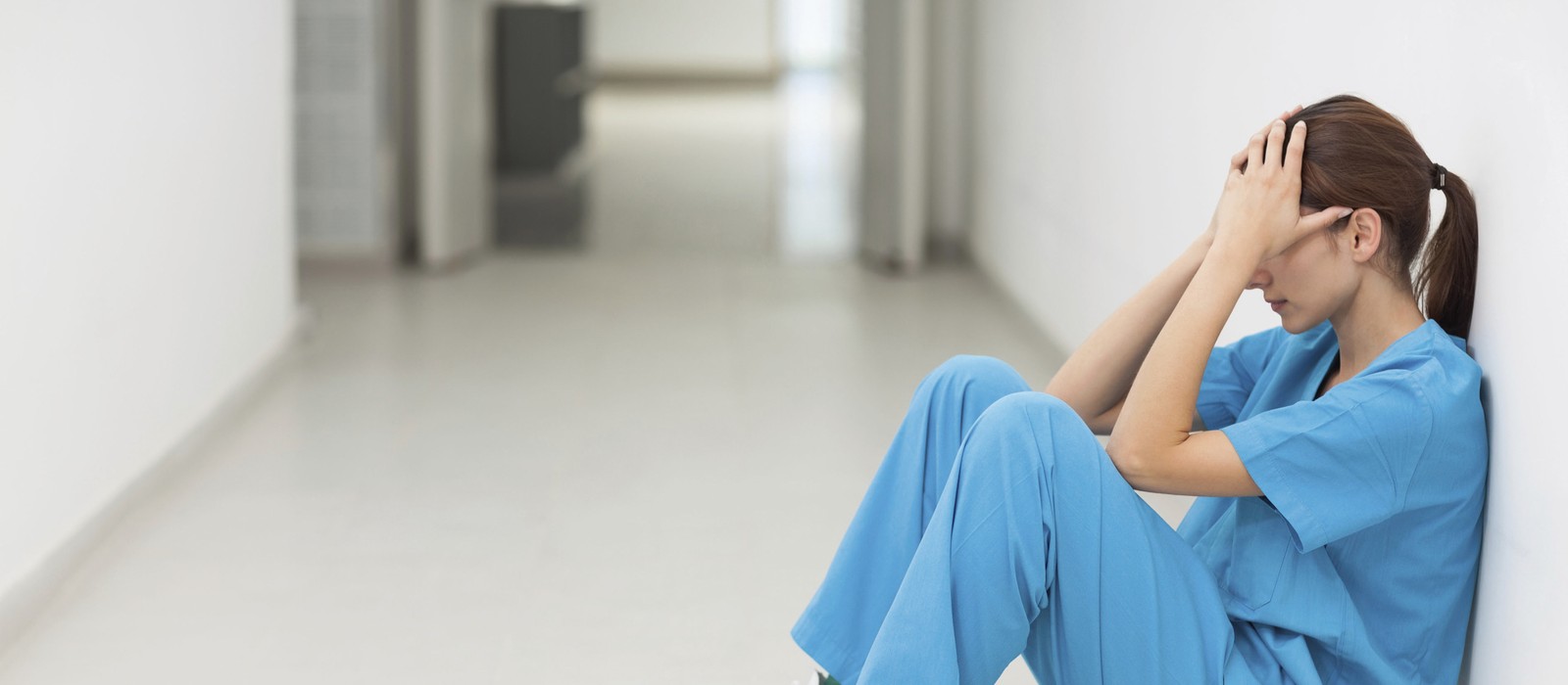 Pflegekraft in blauer Kleidung sitzt erschöpft an einer Wand in einem leeren Krankenhausflur.
