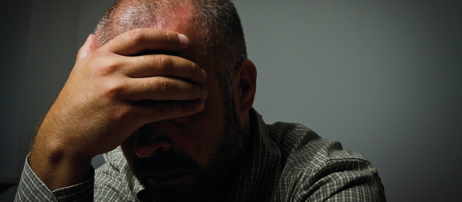 Mann mit gesenktem Kopf und Hand auf der Stirn, in einem dunklen Raum sitzend.