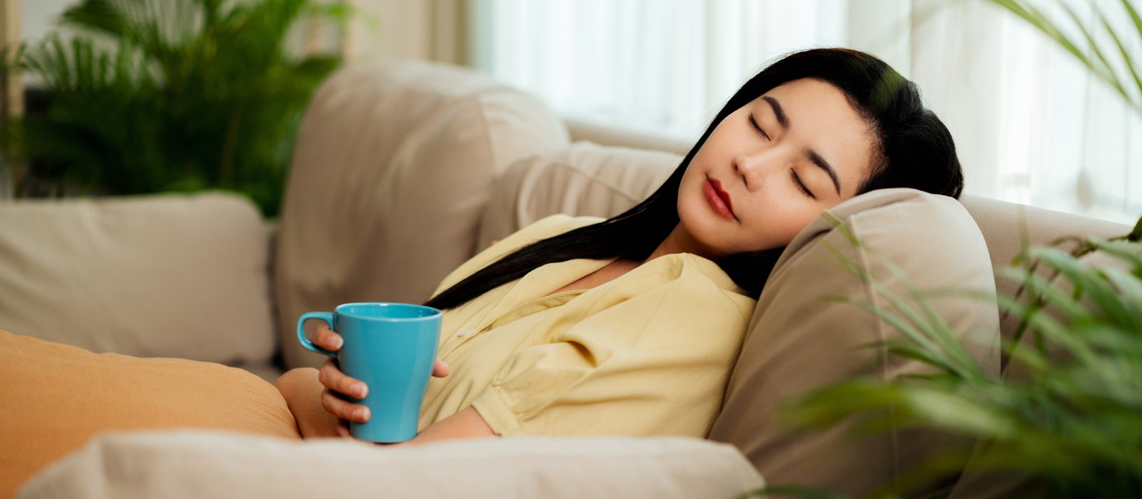 Eine entspannte Person sitzt auf einem Sofa und hält eine Tasse in der Hand, umgeben von Pflanzen.