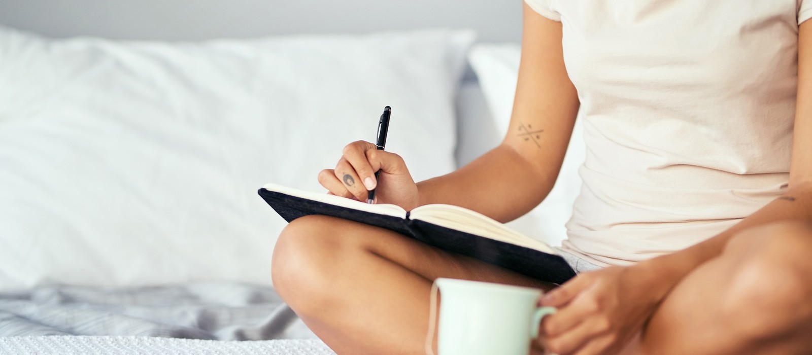 Eine Person sitzt auf dem Bett, schreibt in ein Notizbuch und hält eine Tasse in der Hand.