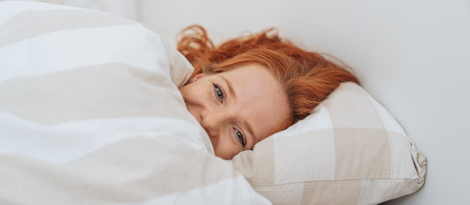 Donna sorridente con capelli rossi, sdraiata a letto sotto una coperta a righe.