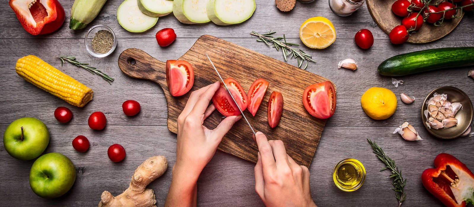 Mani che affettano pomodori freschi su un tagliere di legno, circondati da verdure e spezie.
