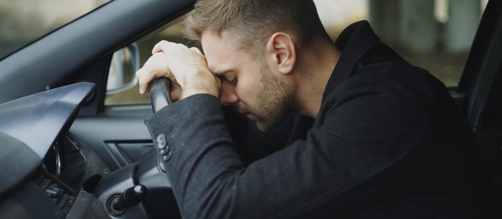 Mann sitzt im Auto mit dem Kopf auf dem Lenkrad, wirkt gestresst und erschöpft.
