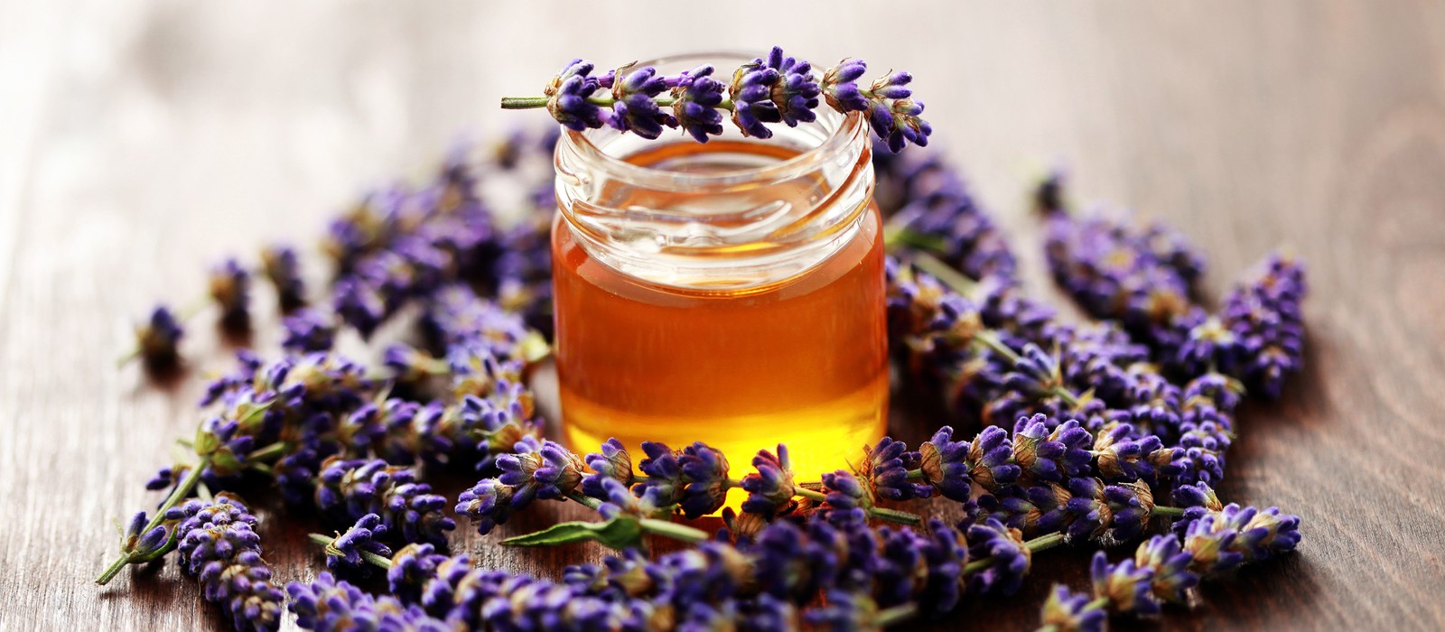 Un bicchiere di miele circondato da fiori di lavanda freschi su un tavolo di legno.