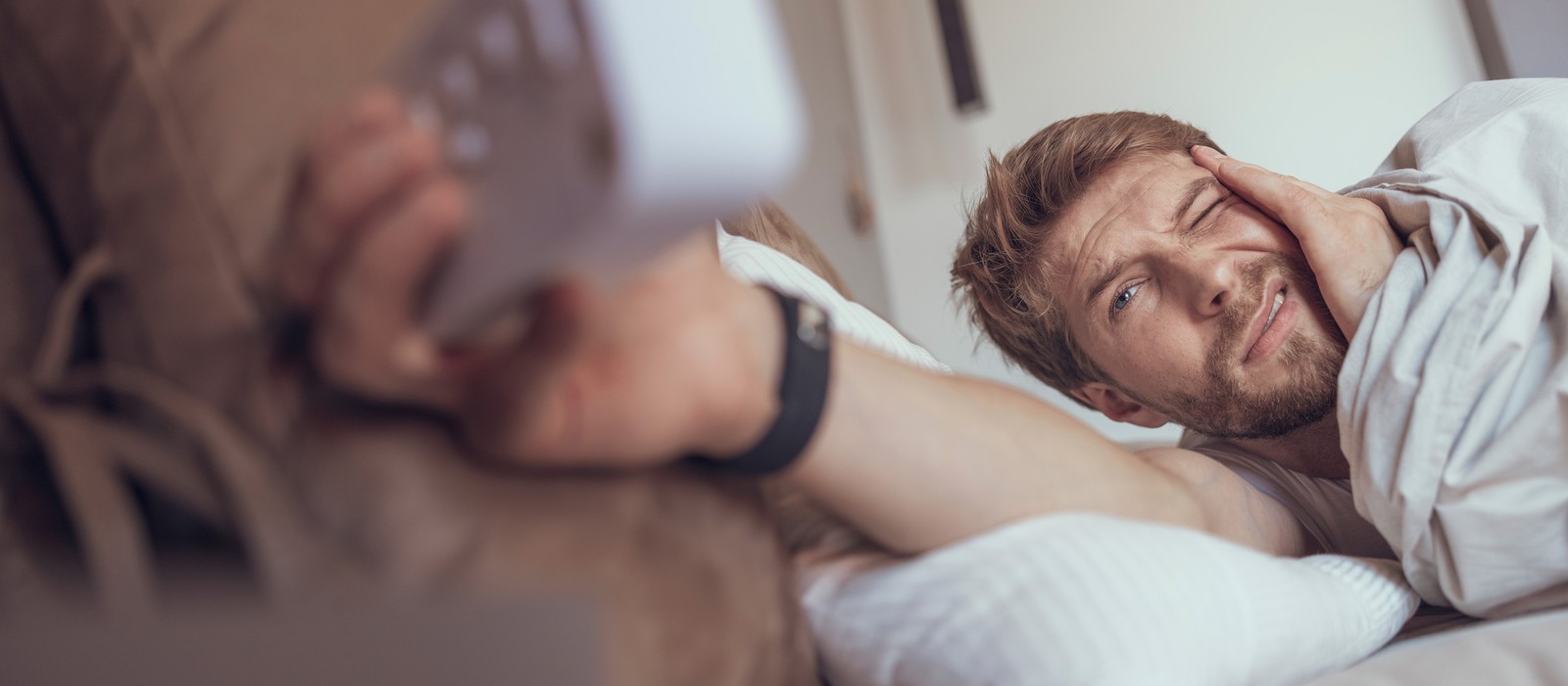 Mann im Bett mit müdem Gesichtsausdruck, hält eine Fernbedienung in der Hand.