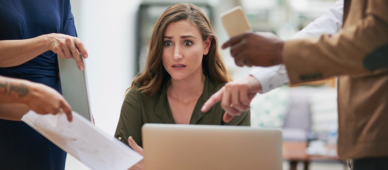 Eine besorgte Frau sitzt vor einem Laptop, umgeben von Personen, die auf Dokumente und Smartphones zeigen.