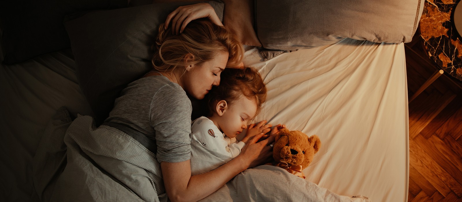 Eine Mutter schläft mit ihrem Kind im Bett, beide umarmt eine Plüschfigur. Sanfte Beleuchtung und gemütliche Bettwäsche.