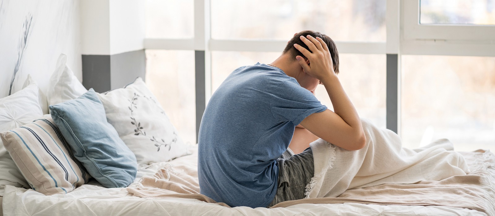 Mann sitzt auf dem Bett, hält den Kopf in den Händen und wirkt nachdenklich oder gestresst.