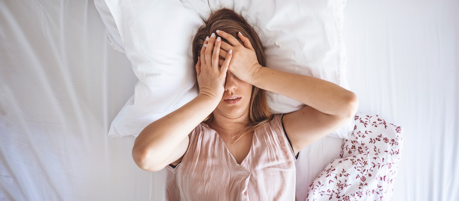 Frau liegt im Bett und hält sich mit beiden Händen das Gesicht, wirkt gestresst oder unruhig.