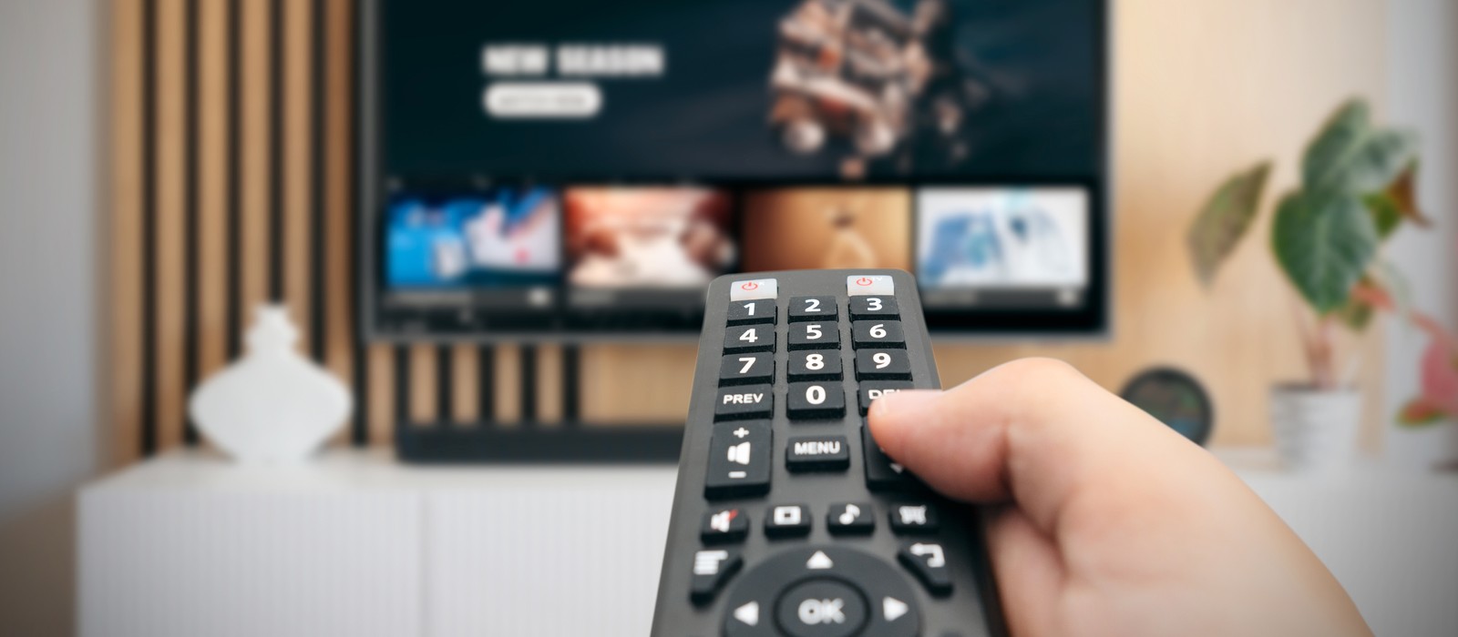 Hand, die eine Fernbedienung hält, während ein Fernseher mit Streaming-Inhalten im Hintergrund zu sehen ist.