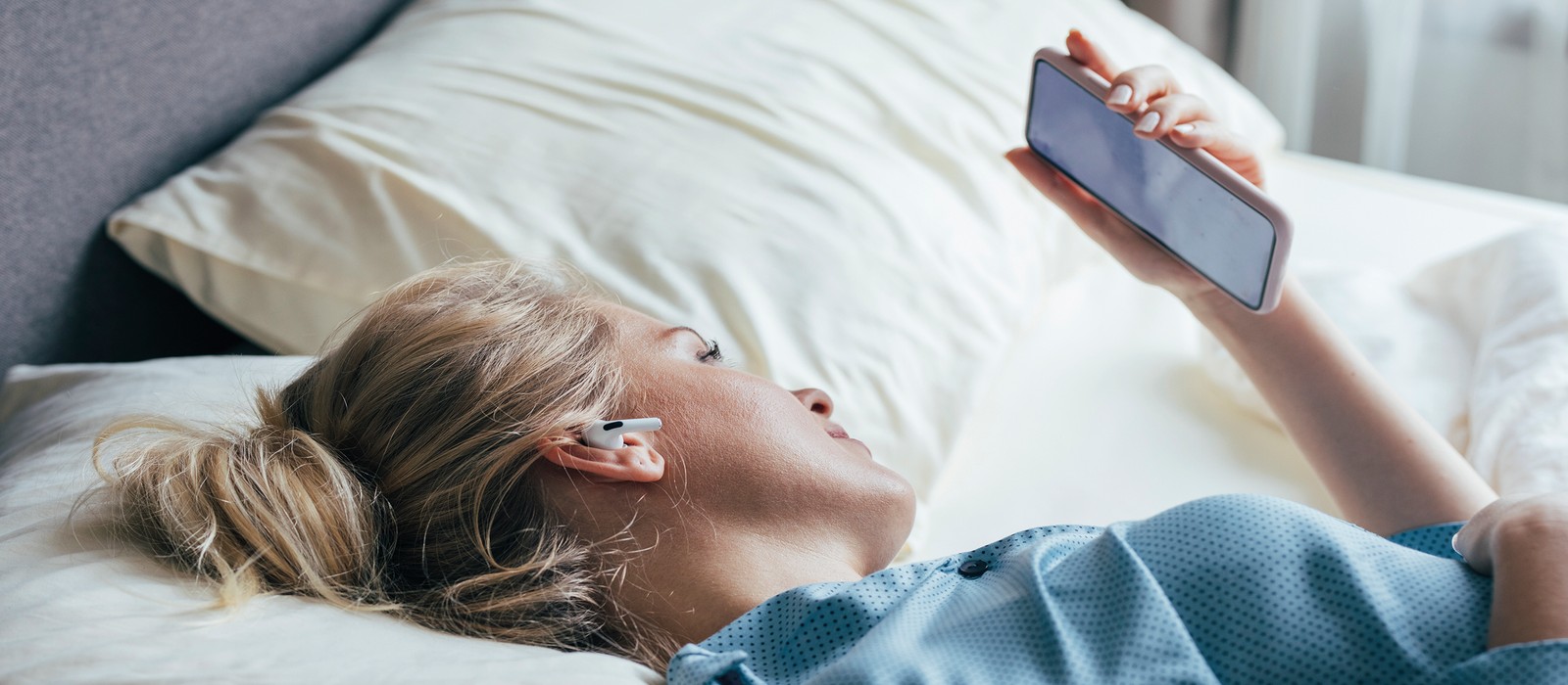Frau liegt im Bett und schaut auf ihr Smartphone, während sie Kopfhörer trägt.