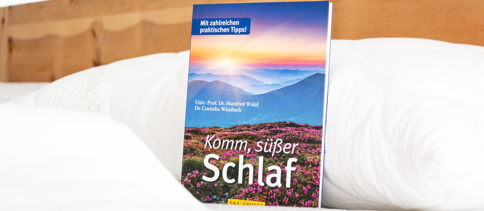 Buchcover mit dem Titel „Komm, süßer Schlaf“ vor einem Bett, umgeben von einer gemütlichen Bettdecke.
