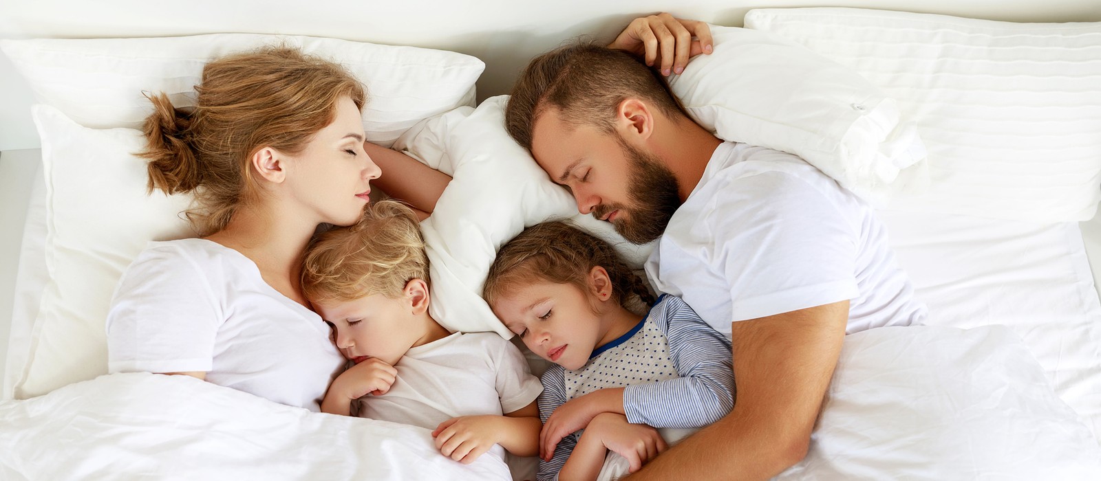 Familie schläft entspannt in einem Bett, umgeben von weißen Kissen und Decken, mit Kindern in der Mitte.