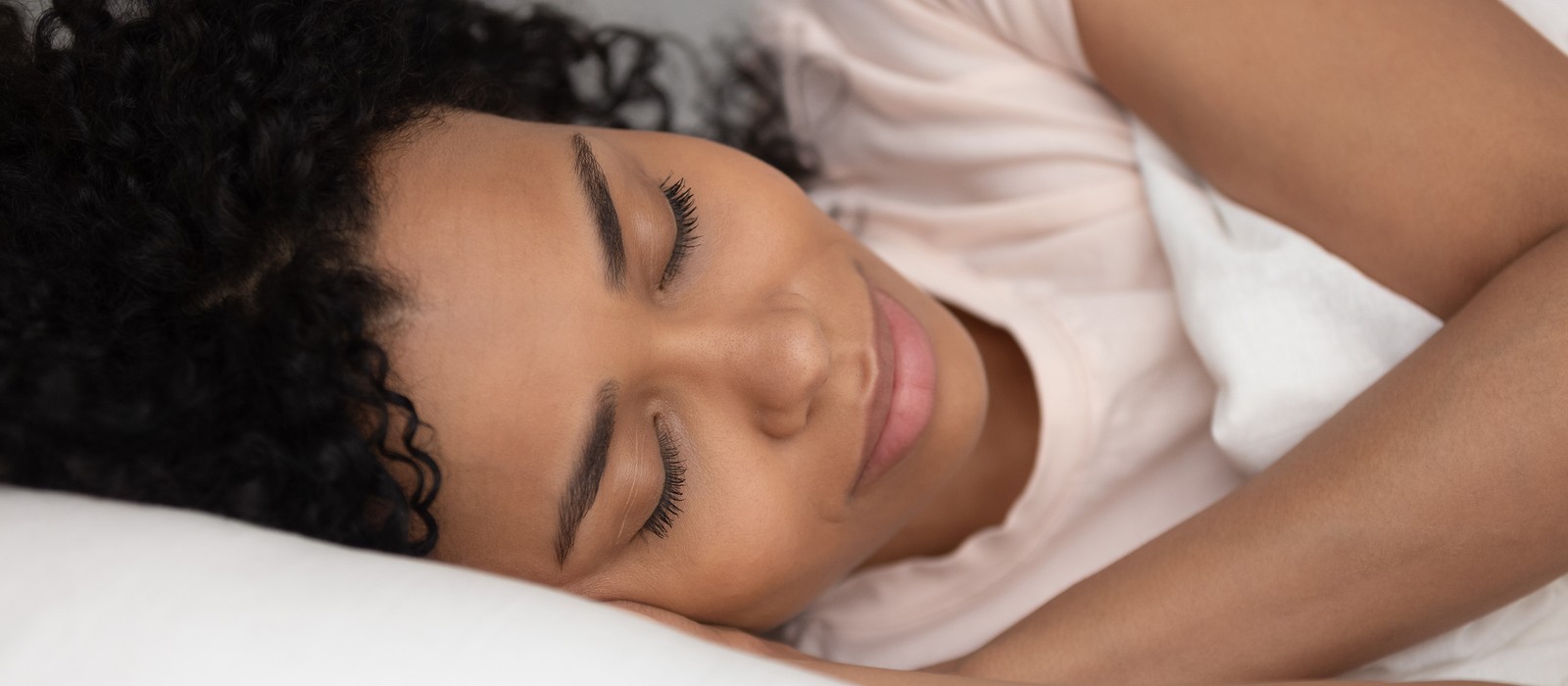 Frau mit lockigem Haar schläft entspannt auf einem Kissen, umgeben von einer Decke.