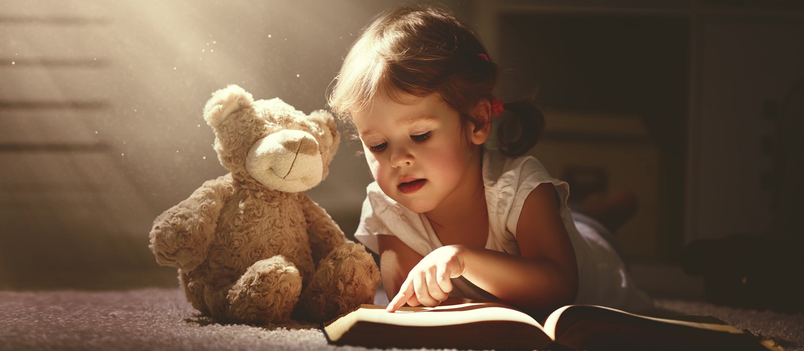 Ein Kind liegt auf dem Boden und liest ein Buch, neben ihm ein Kuscheltier in Form eines Bären.