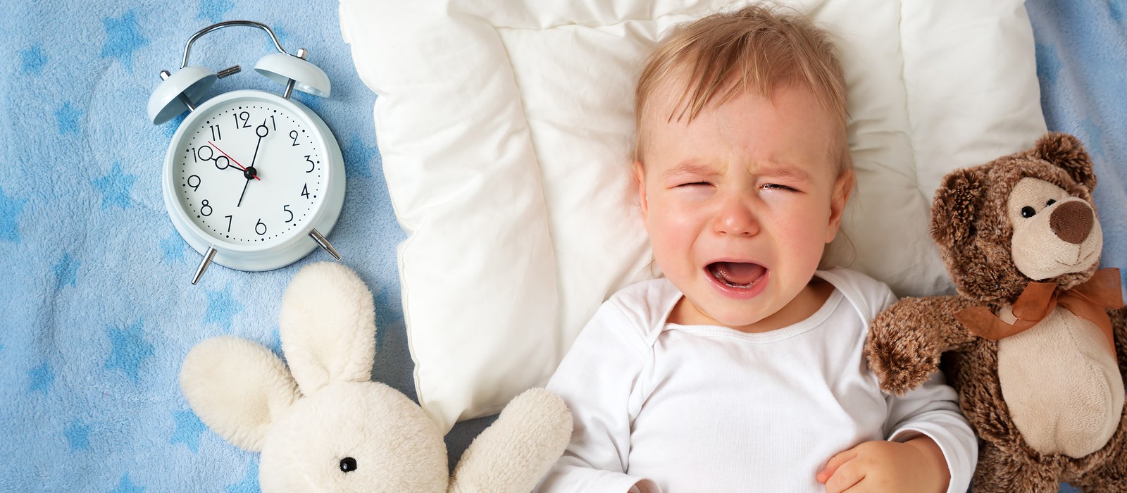 Weinendes Baby auf einem Kissen, umgeben von einem Stoffhasen und einem Bären, mit einer Uhr im Hintergrund.