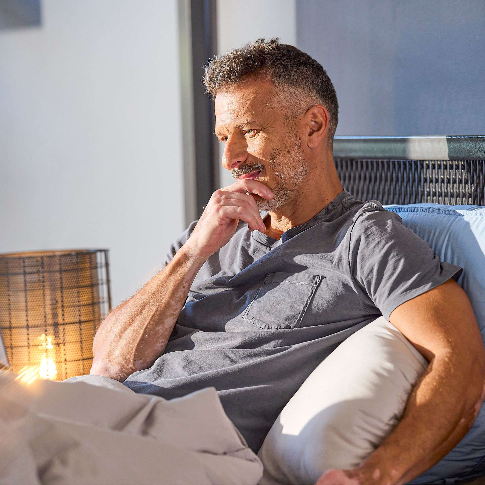 Man sitting relaxed on a bed, thoughtfully resting his chin on his hand, in a bright room.
