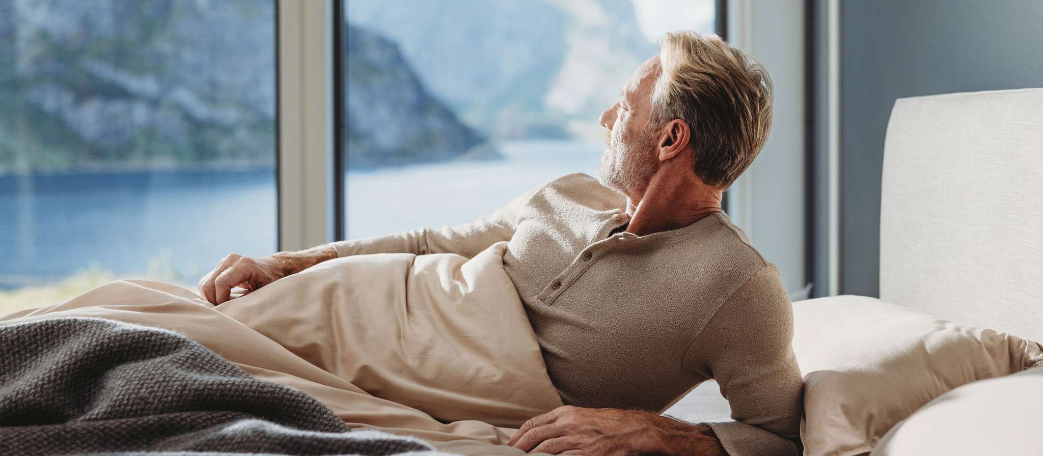 Mann liegt entspannt im Bett und blickt aus dem Fenster auf eine malerische Landschaft.