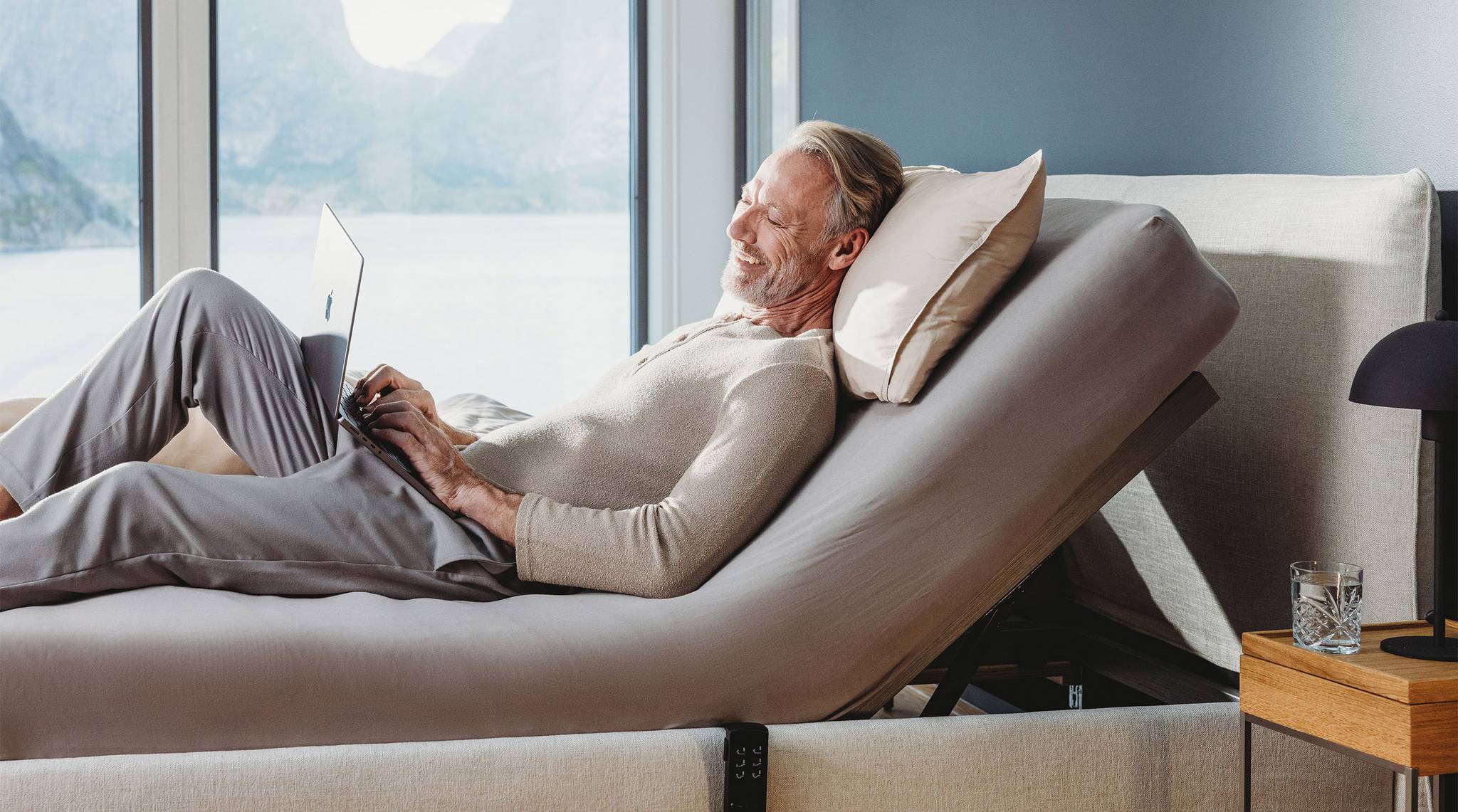 Mann sitzt entspannt auf einem verstellbaren Bett und arbeitet am Laptop mit Blick auf die Landschaft.