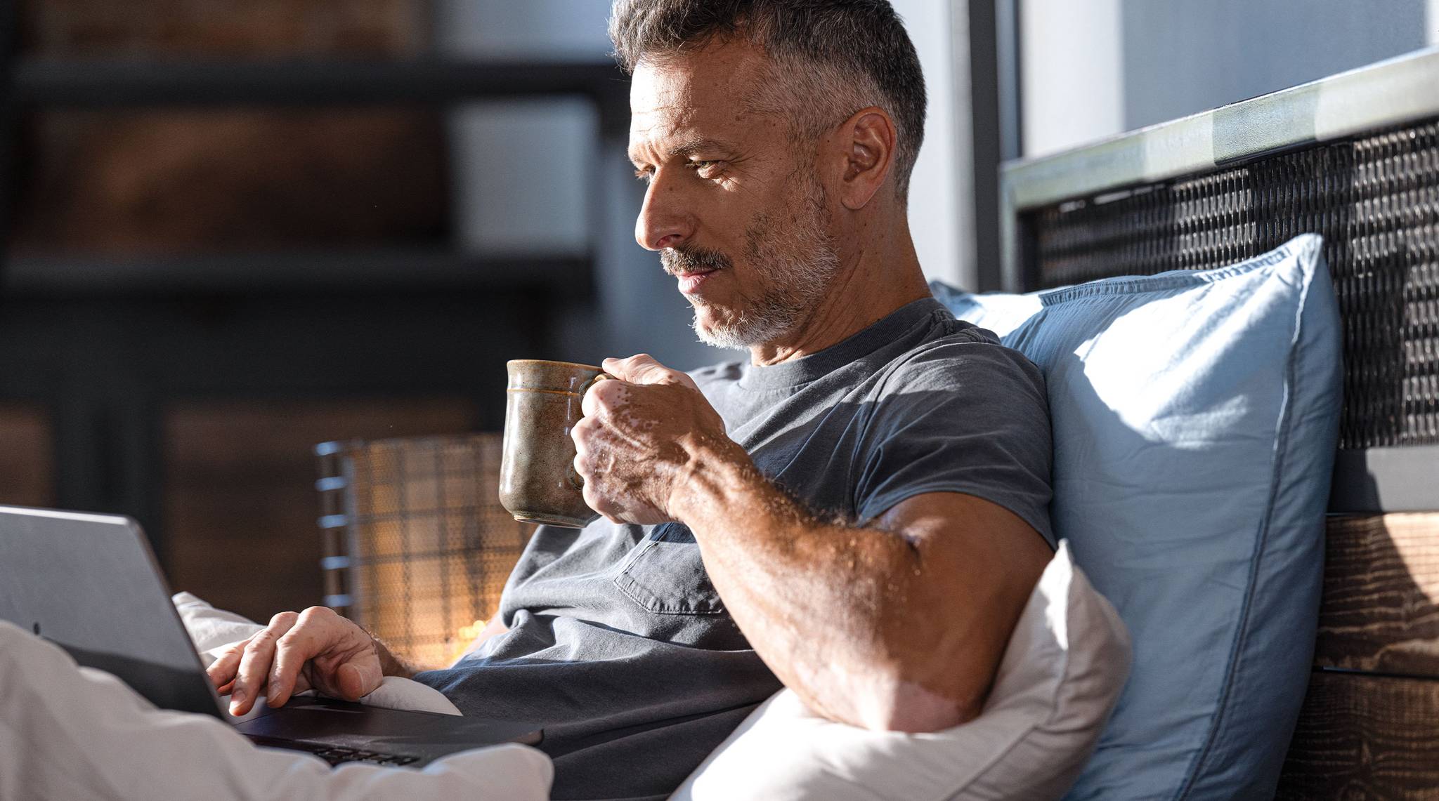 Man sitting relaxed on the bed, drinking from a cup and working on a laptop.