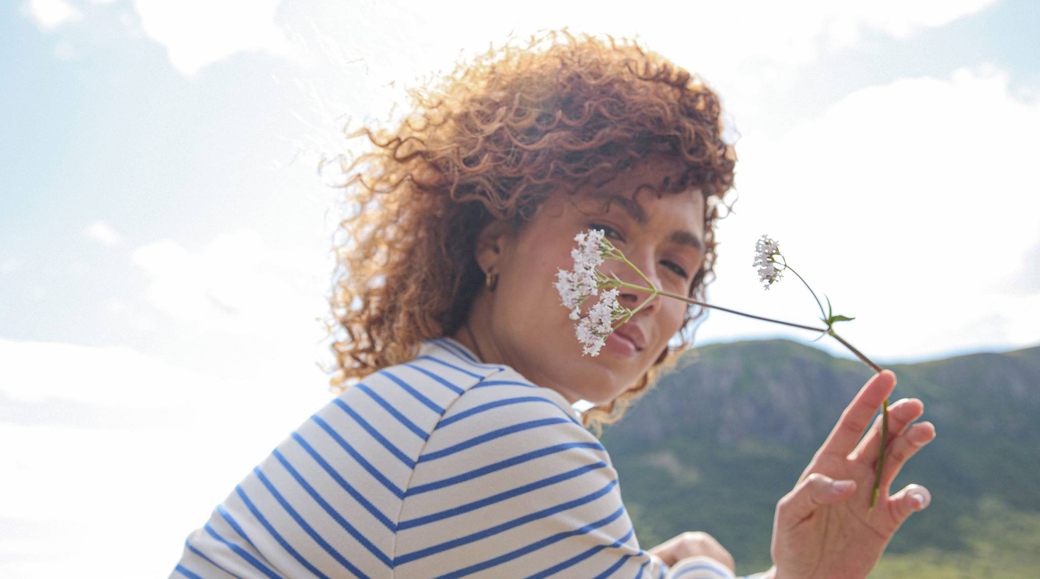 A woman with curly hair holds a flower in front of her face, surrounded by natural landscape.