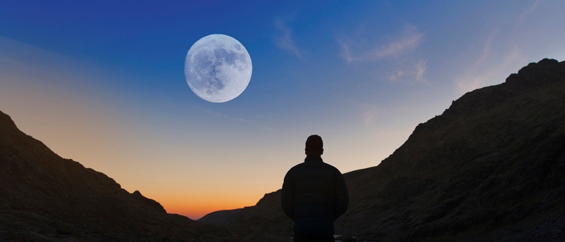 Ein Mann steht in den Bergen bei Sonnenaufgang während der Vollmond am Himmel sichtbar ist