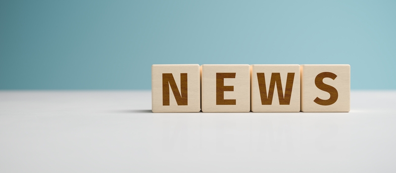 Holzbuchstaben bilden das Wort "NEWS" auf einem hellen Tisch vor einem blauen Hintergrund.