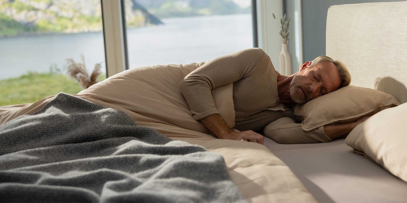 Ein älterer Mann schläft entspannt auf einem Bett mit Kissen, umgeben von einer ruhigen Landschaft.