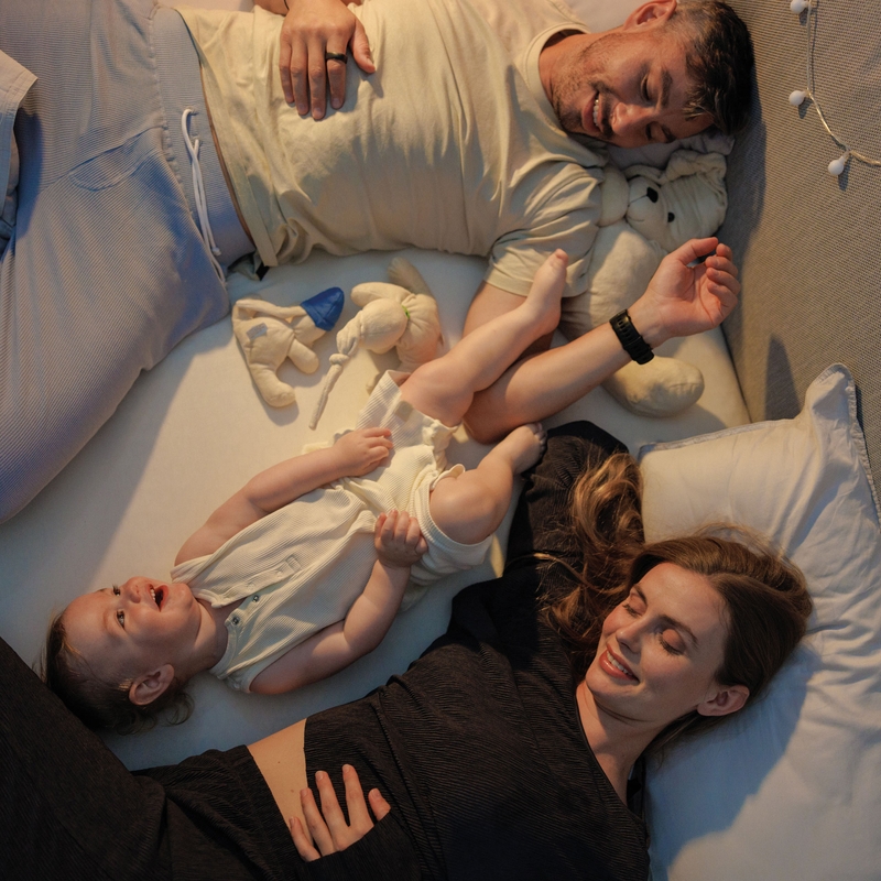 Familie entspannt im Bett mit einem Kleinkind, umgeben von Kissen und Spielzeug, in gemütlicher Schlafumgebung.